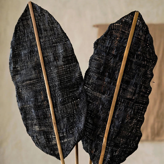 Two large dried black leaves mounted on natural bamboo stakes, displayed against a neutral beige textured wall.