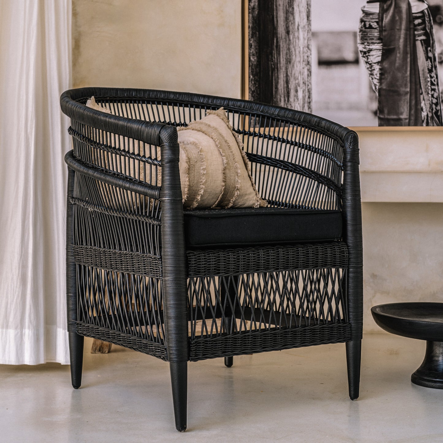 A black rattan chair with a handwoven design, featuring a cushion on the seat, positioned in a room with a light curtain background.