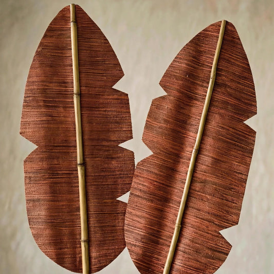 Two decorative banana leaf wall art pieces with a textured reddish-brown pattern and natural bamboo stakes, displayed against a neutral beige wall.