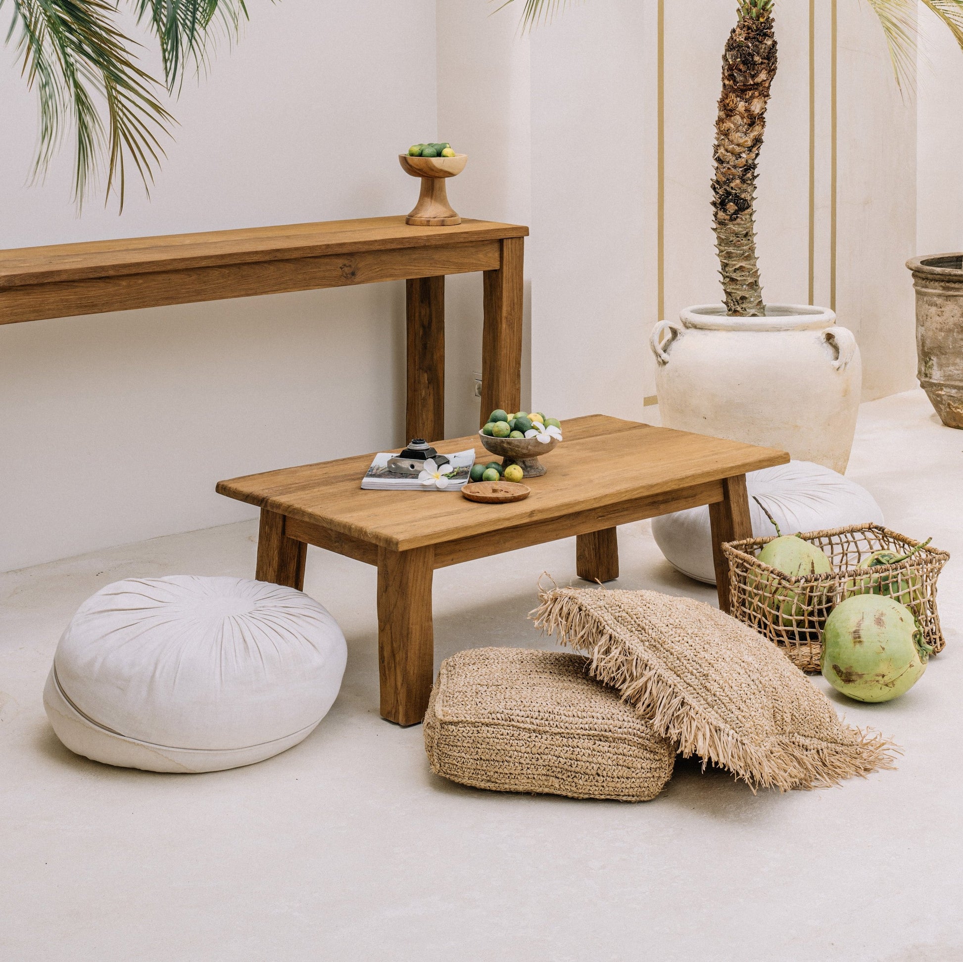 A brown wooden coffee table with a rectangular shape, displayed in a room with palm trees in the background. The table is accompanied by beige cushions and a woven basket, suggesting a casual yet stylish living space.