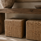 Two woven natural-fiber storage ottomans or baskets sitting underneath a rustic wooden table in a styled interior setting.