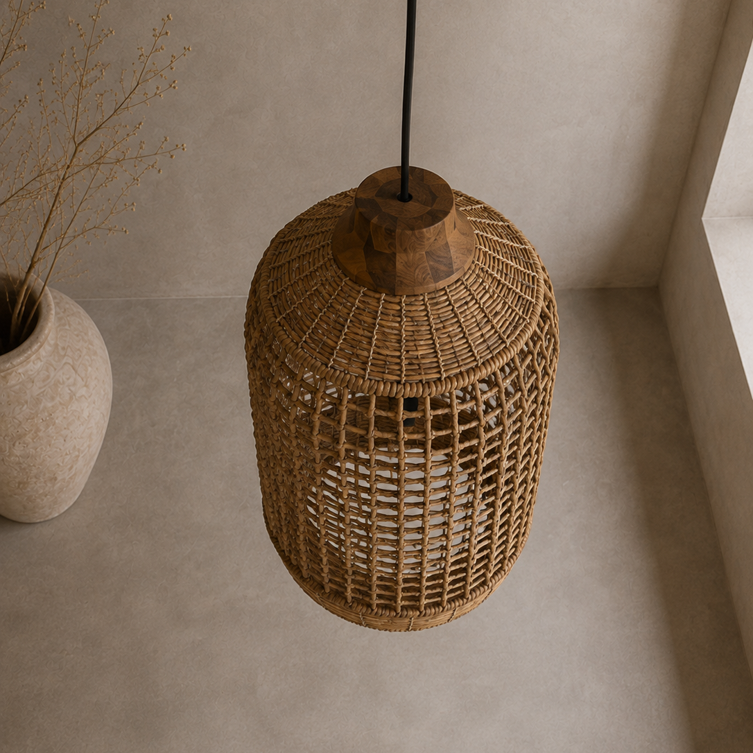 A natural woven rattan pendant light hanging from a black cord in a minimalist room with textured beige walls and floor, next to a ceramic vase with dried branches.