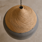 A spherical pendant light with a textured natural woven straw top and a smooth matte gray lower half, suspended by a dark metal chain against a warm beige wall.