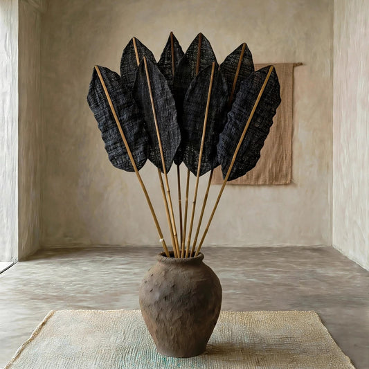 A rustic brown ceramic vase filled with dried black palm leaves on a woven beige rug in a minimalist concrete room.