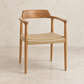 A mid-century modern wooden armchair with a natural oak frame and a woven jute seat, set against a textured beige wall and floor.