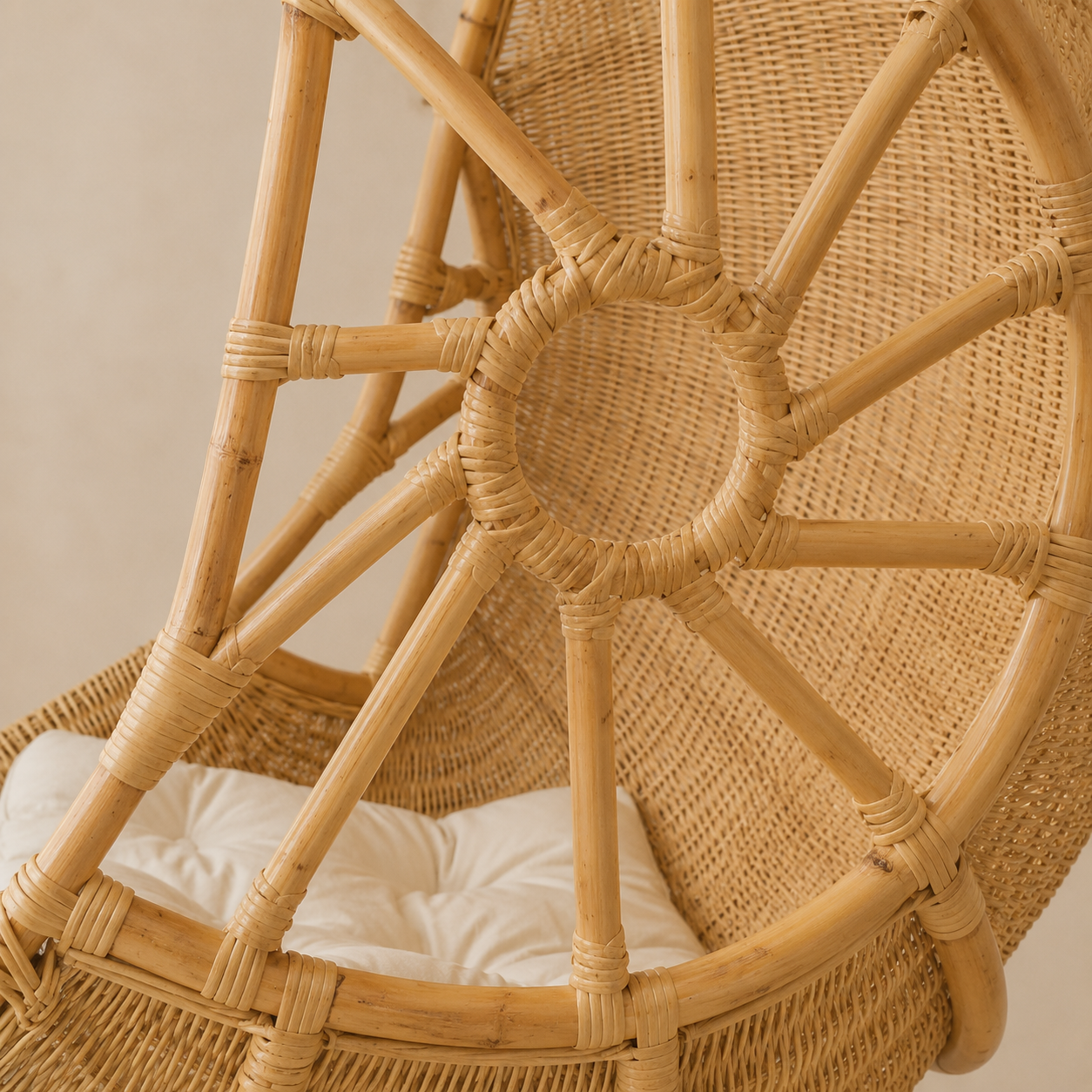A close-up of a natural rattan hanging chair with a white cushion inside, showing the woven circular backrest and radial frame structure.
