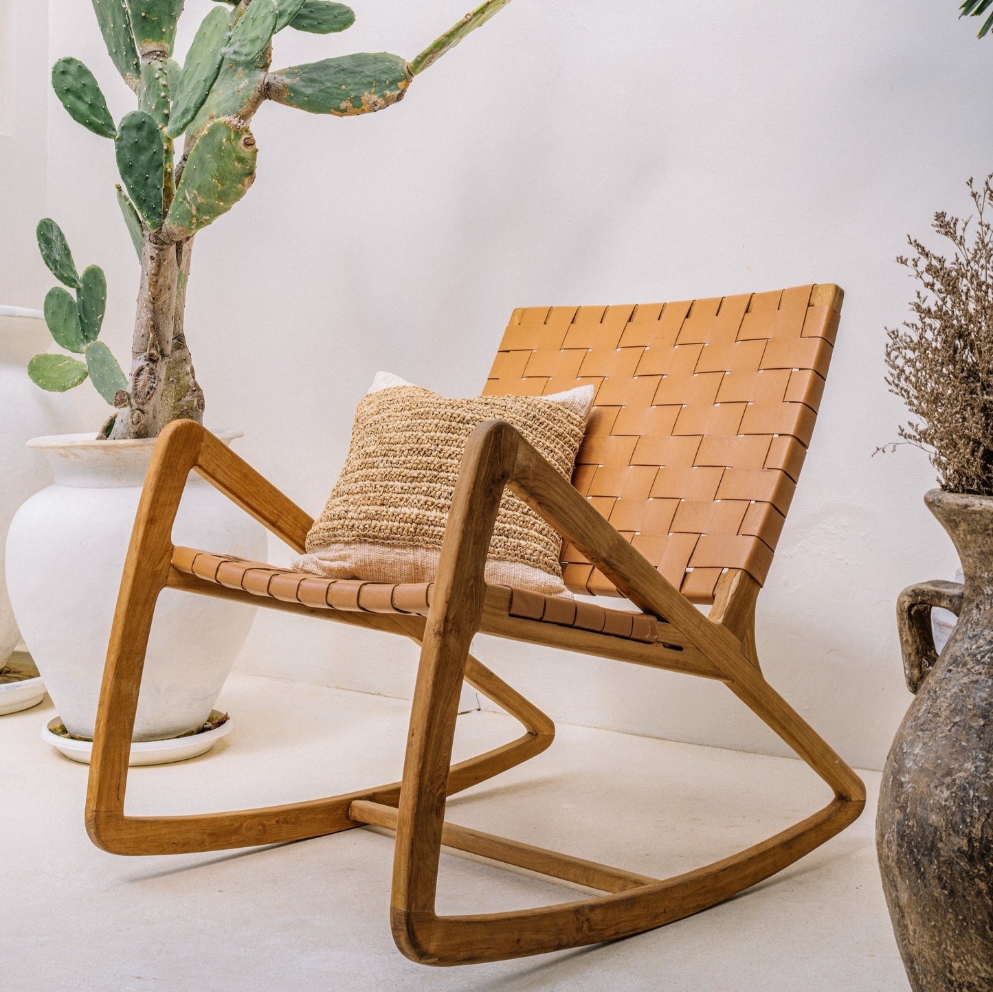 A solid teak wood frame relaxing chair with a leather seat, accompanied by a decorative cushion, positioned in a room with a cactus plant and decorative vases.