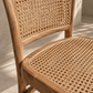 Close-up of a wooden dining chair with a woven cane backrest and seat, shown in warm natural light against a light neutral wall.