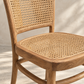 Close-up of a wooden dining chair with a woven cane backrest and seat, set against a textured light beige wall with soft shadow patterns.