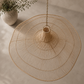 A large bohemian-style woven pendant light made of natural tan raffia or wicker, suspended by a chain from a neutral-toned ceiling in a minimalist room setting with a potted plant in the background.