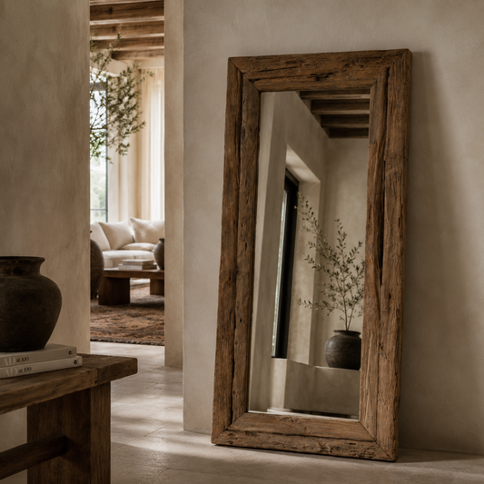 A tall, rustic wooden floor mirror with a thick, weathered brown frame leans against a textured beige plaster wall in a warm, modern interior.