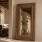 A tall, rustic wooden floor mirror with a thick, weathered brown frame leans against a beige plaster wall in a sunlit room with exposed wooden ceiling beams.