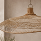 A large woven pendant light made of natural rattan or wicker, shown hanging in a softly lit interior with a warm beige wall and a blurred plant in the background.