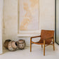 A brown leather chair with teak wood frame, positioned in a room with a light colored wall and decorative artwork, alongside traditional woven baskets.