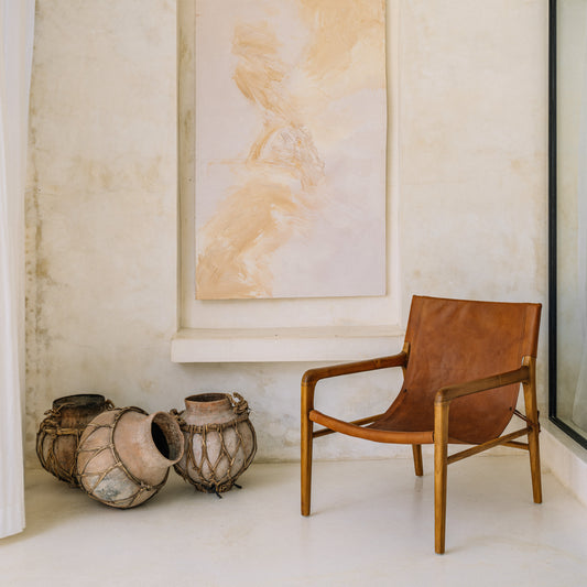 A brown leather chair with teak wood frame, positioned in a room with a light colored wall and decorative artwork, alongside traditional woven baskets.